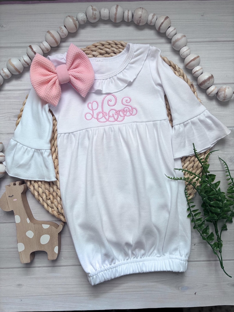May include: A white long-sleeve infant gown featuring a ruffled collar, bell sleeves, and a pink embroidered monogram on the chest. The gown is paired with a matching pink textured fabric bow headband, creating a coordinated set for a baby.