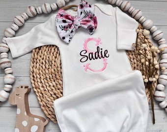 May include: White baby gown with long sleeves and a pink floral bow. The gown has the name "Sadie" embroidered on the front in pink and black.