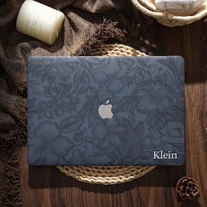 May include: A slate blue laptop case with a floral design, featuring a prominent Apple logo. The case has a textured surface with embossed flowers and leaves. The name "Klein" is printed in white. The case is on a woven mat.