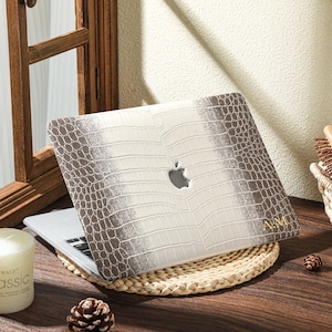 Puede incluir: Un portátil con una funda texturizada con estampado de cocodrilo en tonos blancos y marrones. El logotipo de Apple es visible. El portátil descansa sobre una esterilla tejida, cerca de una vela encendida y una piña.