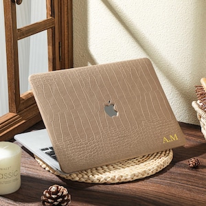 Puede incluir: Una funda para portátil de color marrón claro con textura de piel de cocodrilo, con un recorte para el logotipo de Apple. Las iniciales "A.M" están impresas en dorado. El portátil está sobre una esterilla tejida.