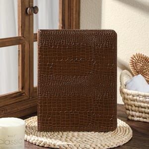 May include: A brown, rectangular journal with a crocodile skin texture. The journal is placed on a woven placemat. A white candle with the text "classic" is in the foreground. A wooden framed mirror is in the background.