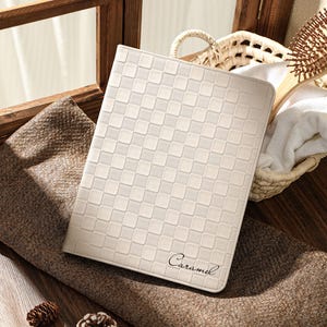 May include: Cream-colored tablet case with a checkered pattern and the word "Caramel" in script. The case is resting on a brown surface near a window. A basket, comb, and pinecones are in the background.