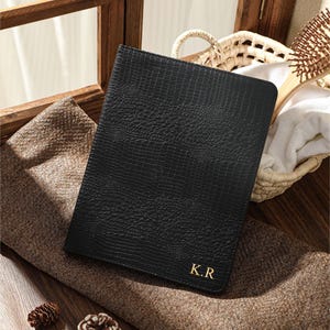 May include: A black rectangular notebook cover with a textured crocodile skin pattern. The bottom right corner features the gold embossed initials K.R. The cover is positioned on a brown fabric surface next to two small pinecones.