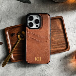 May include: A brown leather phone case with a black frame and a camera cutout. The case has a stitched design and the initials "KH" are embossed in gold on the back.