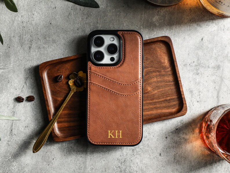 May include: A brown leather phone case with a black frame and a camera cutout. The case has a stitched design and the initials "KH" are embossed in gold on the back.