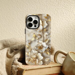 以下が含まれることがあります： 花柄の白いiPhoneケース。金色のアクセントと大理石の背景が特徴です。