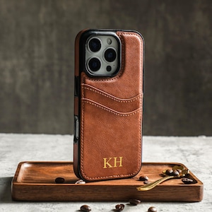 May include: A brown leather phone case with a card slot and a black camera cutout. The case has the initials "KH" embossed in gold on the back.