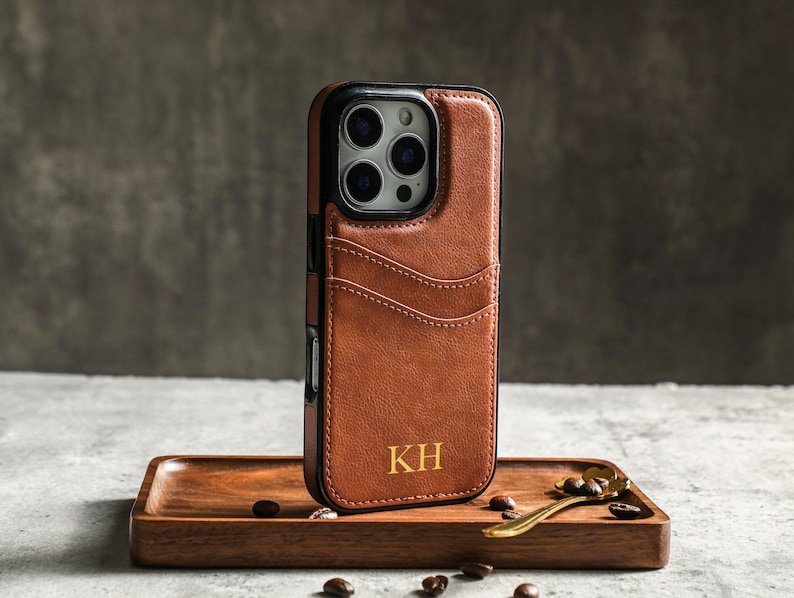 May include: A brown leather phone case with a card slot and a black camera cutout. The case has the initials "KH" embossed in gold on the back.
