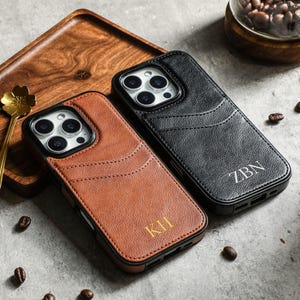 May include: Two leather phone cases with a wallet feature. The case on the left is brown with the initials "KH" embossed on the back. The case on the right is black with the initials "ZBN" embossed on the back.