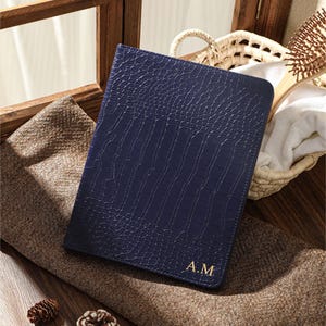 May include: A rectangular navy blue notebook with a textured crocodile skin pattern on the cover. The bottom right corner features the gold embossed initials A.M. The notebook has rounded corners and a slim, vertical orientation.