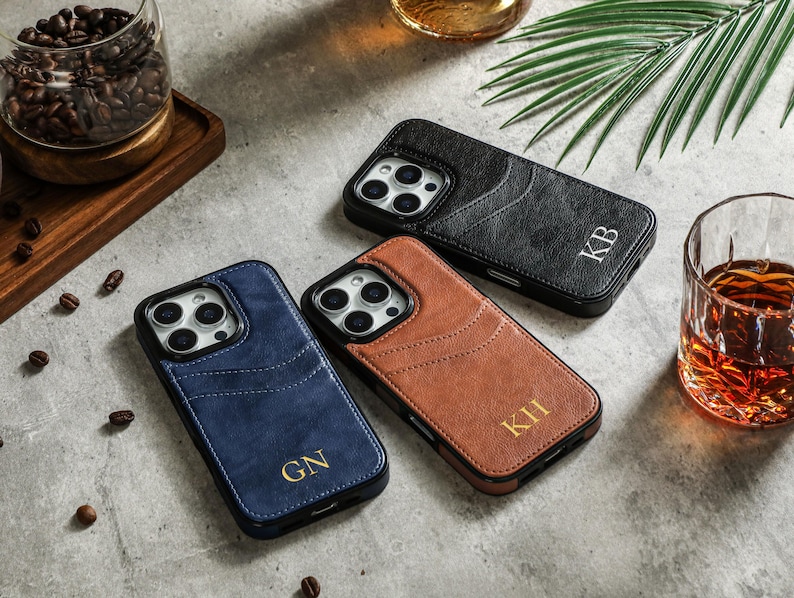 May include: Three different colored leather phone cases with personalized initials. The cases are black, brown, and blue. The initials are "KB", "KII", and "GN".
