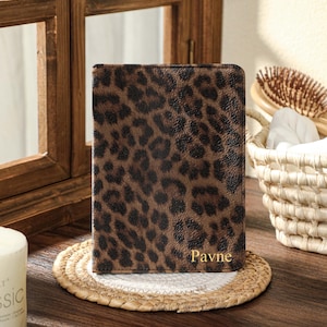 May include: A brown and black leopard print journal with the name "Pavne" in gold lettering. The journal is on a round woven coaster. The background includes a wooden window frame, a candle, and a basket.