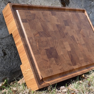 May include: A large, rectangular wooden cutting board with a thick, raised edge. The board is made of multiple pieces of wood, creating a patchwork pattern. The wood is a rich brown color with a natural grain.