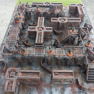 Peut inclure: Une maquette détaillée d'une ville futuriste avec plusieurs niveaux et passerelles. La maquette est en métal et peinte dans une couleur brun rouille. De petites figurines de personnes et de véhicules sont dispersées dans toute la maquette.