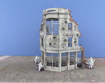 May include: A miniature, weathered building with a corner design, featuring multiple windows and visible bullet holes. The structure has a partially destroyed facade with exposed brick. Small soldier figurines are positioned around the building, suggesting a war scene.