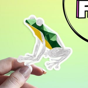 Peut inclure: Un autocollant représentant une grenouille géométrique. La grenouille est principalement verte avec des accents jaunes, soulignée de blanc, et a une apparence stylisée et détaillée. L'autocollant est tenu par une main sur un fond vert dégradé.