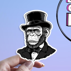 Op de afbeelding: Een zwart-witte sticker met een chimpansee die een hoge hoed, een pak en een vlinderdas draagt. De chimpansee heeft een serieuze uitdrukking. De sticker staat op een lichtblauwe achtergrond. De sticker wordt door een hand vastgehouden.