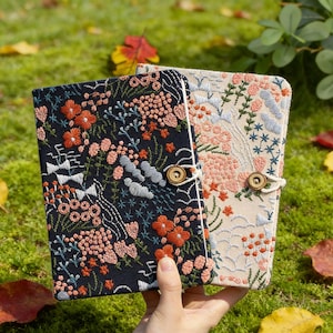 Op de afbeelding: Twee geborduurde notitieboekjes met bloemmotieven. Eén notitieboekje heeft een marineblauwe kaft met oranje en roze bloemen, de andere heeft een crèmekleurige kaft met roze en blauwe bloemen. Beide hebben een houten knoopsluiting.