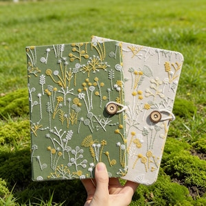 May include: Two embroidered journals with floral designs. One is olive green, the other is beige. Both have a wooden button closure and cream string. The floral embroidery includes white and yellow accents.