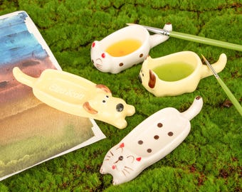 Pode incluir: Quatro paletas de tinta de cerâmica em forma de animais: um gato e dois cães. A paleta de gato é branca com manchas marrons. As paletas de cães são amarelas. As paletas estão em uma superfície verde com pincéis.