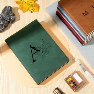 Op de afbeelding: Een donkergroen notitieboek met de letter "A" en een bloemmotief wordt getoond. Een stapel notitieboeken met verschillende gekleurde ruggen is zichtbaar. Een aquarelpalet en een penseel zijn ook aanwezig, wat wijst op een briefpapier thema.