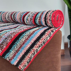 May include: A rolled-up, colorful crochet blanket with a red border. The blanket features stripes of black, red, brown, blue, and white. The blanket is resting on a brown surface.