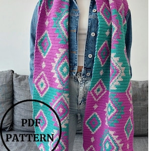 Op de afbeelding: Een gehaakte sjaal met een geometrisch patroon in tinten roze, turquoise en wit. De sjaal heeft franjes aan de uiteinden en wordt gedragen door iemand die een spijkerjack en jeans draagt. De tekst "PDF PATTERN" is zichtbaar in de afbeelding.