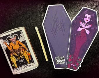 May include: An image featuring a matchbox with "THE DEVIL" tarot card art, a match, and a coffin-shaped sticker with a vampire illustration. The sticker has the text "BATRIDER DESIGNS" on it. The items are on a black surface.