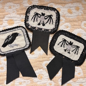 May include: Three rectangular fabric badges with black scalloped lace borders. One badge features an embroidered black crow, while the other two display embroidered silver and black bats. Each badge has a black ribbon tail attached at the bottom.