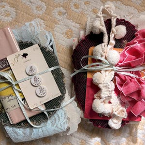 May include: Two bundles of sewing supplies tied with light blue ribbon. The left bundle includes pink rayon hem facing, light blue lace, and three white buttons with printed designs. The right bundle features dark red fabric, cream pom-pom trim, and pink ruffled fabric.