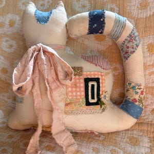May include: A stuffed fabric cat figure in off-white, featuring a long tail decorated with various blue and floral patterned fabric patches. The cat has a large, frayed pale pink ribbon tied around its neck and a rectangular patch on its side.