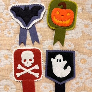 May include: Four felt Halloween-themed badges with scalloped edges. One features a black bat on a blue background, one an orange jack-o'-lantern on green, one a white skull and crossbones on red, and one a white ghost on a black background.