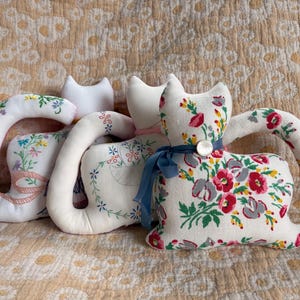 May include: Three handmade cat-shaped pillows with floral and embroidered designs. The pillows feature white fabric with red, pink, and green floral patterns. One cat has a blue ribbon and button detail.