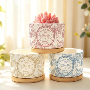 Peut inclure: Trois pots de fleurs cylindriques en céramique blanche avec soucoupes en bambou, ornés d'un motif floral et d'oiseaux au trait. Chaque pot présente un emblème circulaire central montrant deux mains tenant des bagues, des noms et des dates.