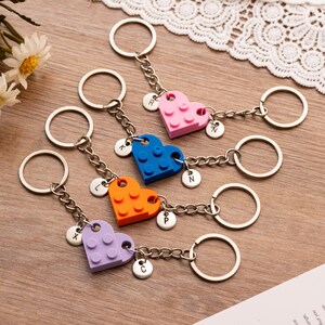 Peut inclure: Cinq porte-clés avec des breloques en forme de cœur faites de blocs de construction colorés. Chaque porte-clés a une chaîne argentée, un charm rond avec une lettre et un anneau métallique. Les blocs sont roses, bleus, oranges et violets.