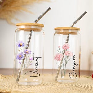 Puede incluir: Dos vasos de vidrio transparente con tapas de bamb&uacute; y pajitas de metal. Uno presenta flores moradas y el nombre "Vincent", el otro tiene flores rosas y el nombre "Grayson". Ambos est&aacute;n sobre una estera tejida.