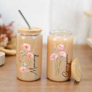 Puede incluir: Dos vasos de vidrio para bebidas con tapas de bamb&uacute;. Un vaso tiene una pajita de acero inoxidable. Ambos vasos presentan dise&ntilde;os florales y los nombres "Mommy" y "Grayson". Los vasos est&aacute;n llenos de un l&iacute;quido claro.
