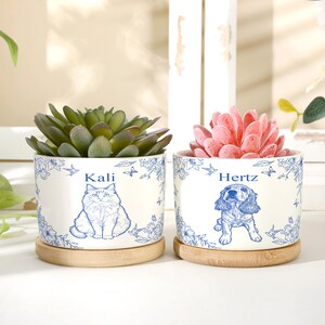Op de afbeelding: Twee cilindrische plantenbakken van wit keramiek met blauwe bloemmotieven en bamboe onderzetters. De ene bak toont een kattenillustratie met de naam Kali, de andere een hondenillustratie met de naam Hertz, beide gevuld met een vetplant.