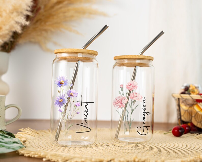 Puede incluir: Dos vasos de vidrio transparente con tapas de bamb&uacute; y pajitas de metal. Uno presenta flores moradas y el nombre "Vincent", el otro tiene flores rosas y el nombre "Grayson". Ambos est&aacute;n sobre una estera tejida.