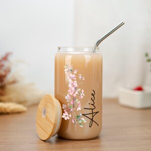 Puede incluir: Un vaso transparente en forma de lata, con tapa de bamb&uacute; y pajita de acero inoxidable. Presenta un dise&ntilde;o floral en rosa y verde, y el nombre "Alice" en escritura negra. El vaso contiene una bebida de color claro.