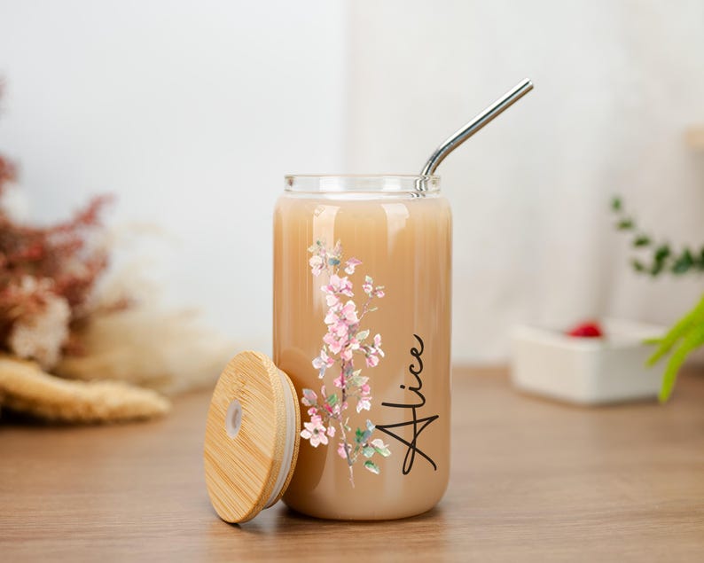 Puede incluir: Un vaso transparente en forma de lata, con tapa de bamb&uacute; y pajita de acero inoxidable. Presenta un dise&ntilde;o floral en rosa y verde, y el nombre "Alice" en escritura negra. El vaso contiene una bebida de color claro.