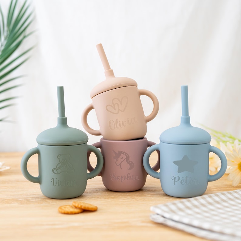 May include: Four pastel-colored silicone sippy cups with handles and straws. Each cup is a different color: green, pink, peach, and blue. The cups are engraved with names and designs, including a star, unicorn, and hearts.