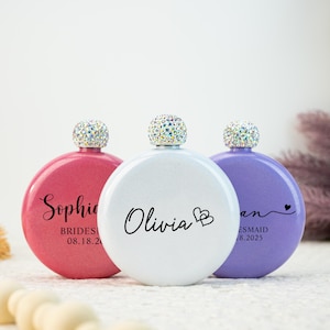 Pode incluir: Três frascos redondos em rosa, branco e roxo, cada um com uma tampa brilhante com joias. O frasco rosa tem o nome "Sophie" e uma data, o branco "Olivia" e corações, e o roxo "an" e uma data.
