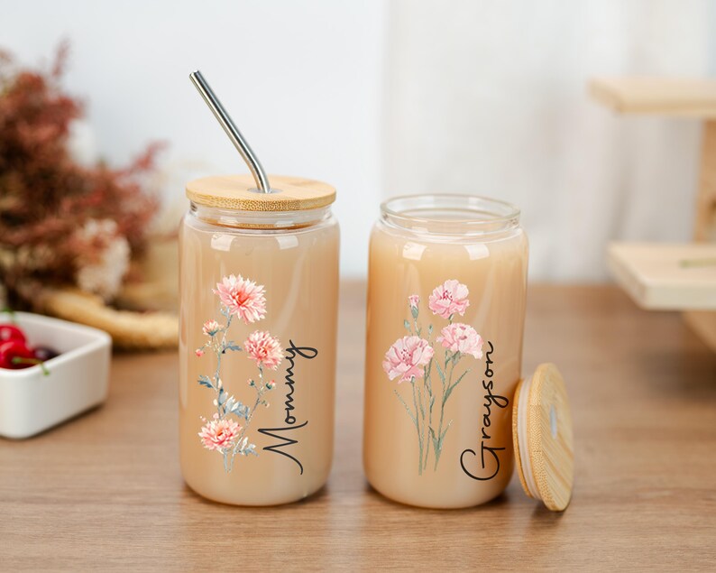 Puede incluir: Dos vasos de vidrio para bebidas con tapas de bamb&uacute;. Un vaso tiene una pajita de acero inoxidable. Ambos vasos presentan dise&ntilde;os florales y los nombres "Mommy" y "Grayson". Los vasos est&aacute;n llenos de un l&iacute;quido claro.