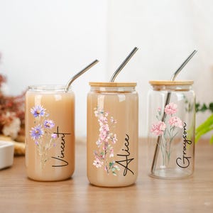 Puede incluir: Tres vasos de vidrio con tapas de madera y pajitas de acero inoxidable. Cada vaso presenta un dise&ntilde;o floral y un nombre: Vincent, Alice y Grayson. Los vasos est&aacute;n llenos de una bebida de color claro y est&aacute;n sobre una superficie de madera.