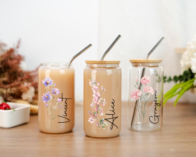 Puede incluir: Tres vasos de vidrio con tapas de madera y pajitas de acero inoxidable. Cada vaso presenta un dise&ntilde;o floral y un nombre: Vincent, Alice y Grayson. Los vasos est&aacute;n llenos de una bebida de color claro y est&aacute;n sobre una superficie de madera.