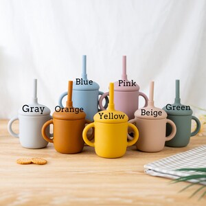 May include: A collection of colorful silicone cups with handles and straws. The cups are in various colors, including gray, orange, blue, yellow, pink, beige, and green. Each cup has a matching lid and straw, with the color names printed above each cup.