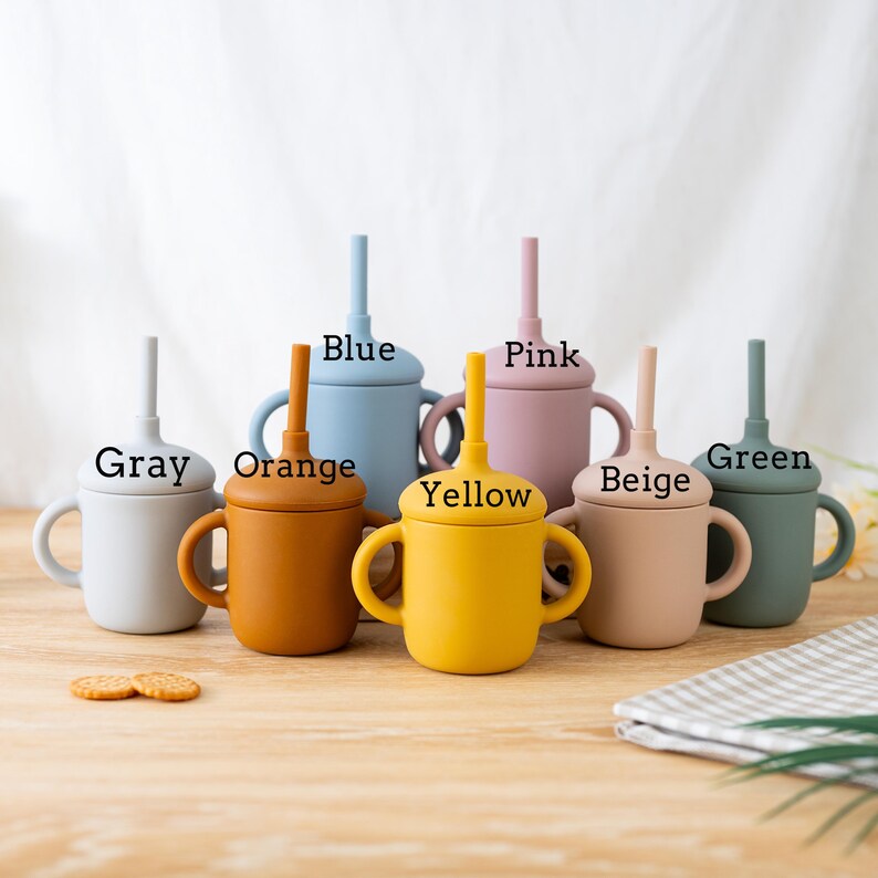May include: A collection of colorful silicone cups with handles and straws. The cups are in various colors, including gray, orange, blue, yellow, pink, beige, and green. Each cup has a matching lid and straw, with the color names printed above each cup.