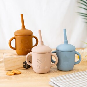 May include: Three silicone sippy cups in shades of brown, pink, and blue, each with a lid and straw. The cups have handles and are personalized with names and designs. The brown cup says "Helen", the pink cup says "Olivia", and the blue cup says "Peter".